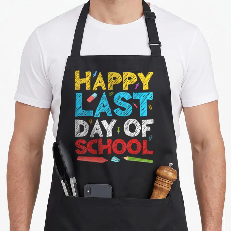 Funny school teacher apron - HAPPY LAST DAY OF SCHOOL text with eraser sharpener pencil and push pins — shown on a black cotton canvas apron against a white background