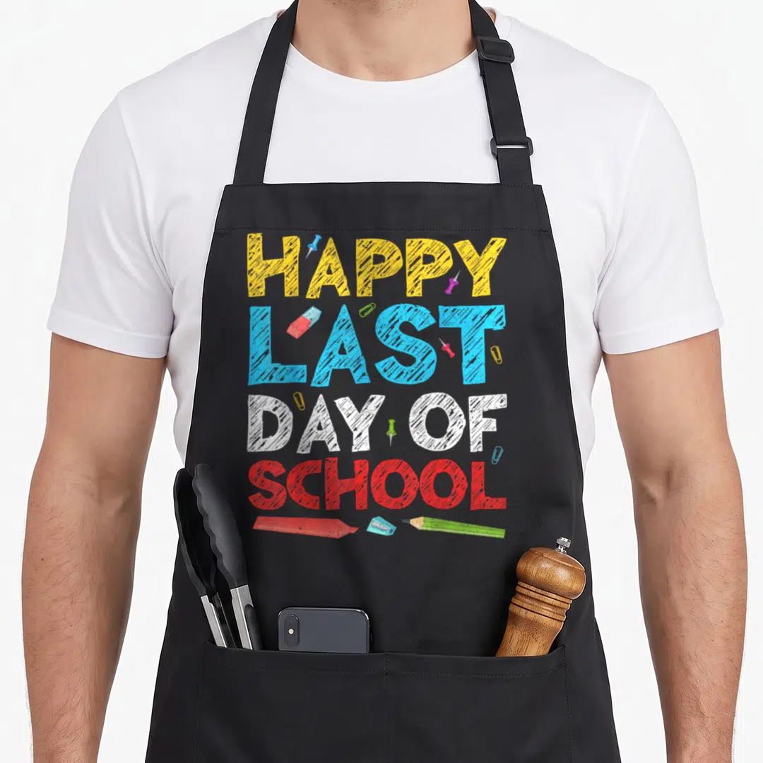 Funny school teacher apron - HAPPY LAST DAY OF SCHOOL text with eraser sharpener pencil and push pins — shown on a black cotton canvas apron against a white background