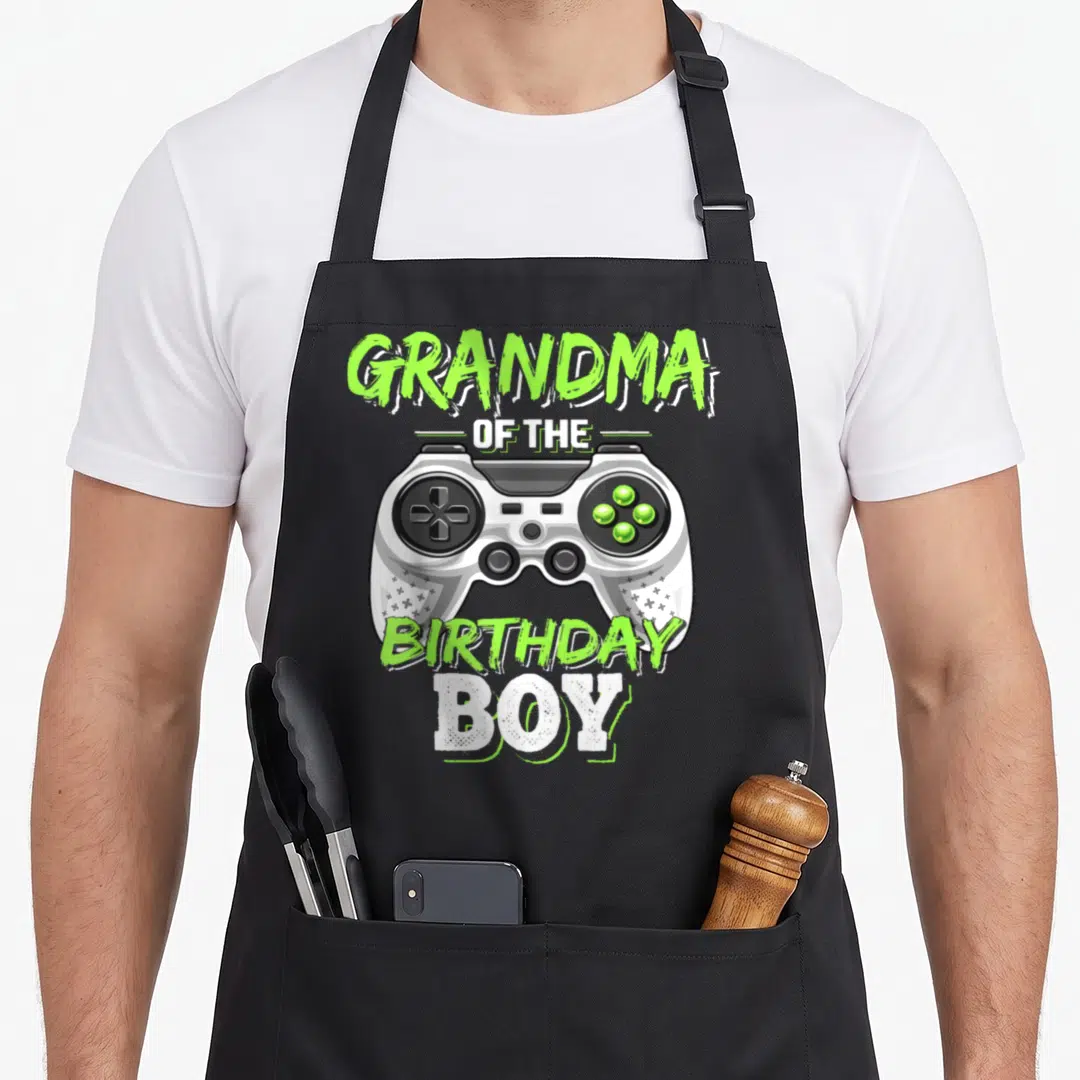 Birthday grandma in apron - Lime green text reading Grandma of the Birthday Boy surrounding a grey game controller — shown on a black cotton canvas apron against a white background