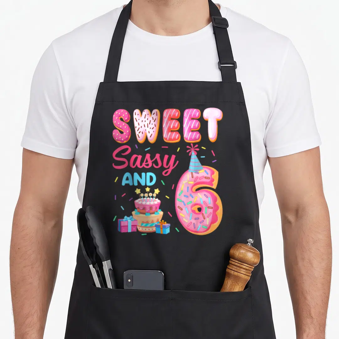 Donut sassy apron - Donut number 6 with party hat, cake, two gift boxes, and sprinkles in pink and teal — shown on a black cotton canvas apron against a white background