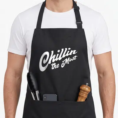 Chillin potato apron - White script text Chillin the Most stacked vertically on grey background — shown on a black cotton canvas apron against a white background
