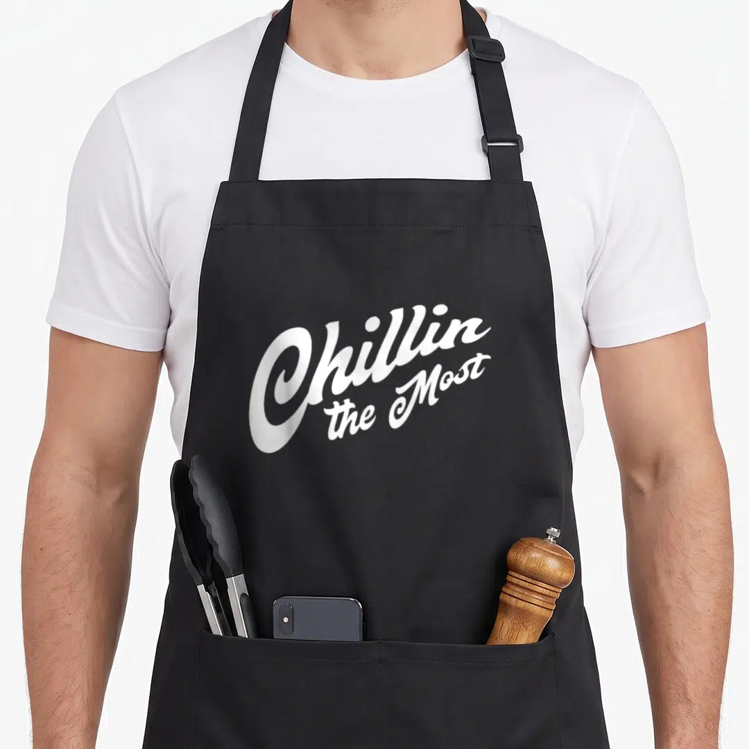 Chillin potato apron - White script text Chillin the Most stacked vertically on grey background — shown on a black cotton canvas apron against a white background