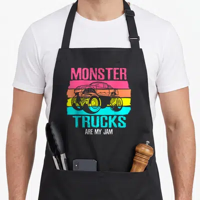 Jam engine truck apron - Retro monster truck on pink orange yellow and cyan stripes with text MONSTER TRUCKS ARE MY JAM — shown on a black cotton canvas apron against a white background