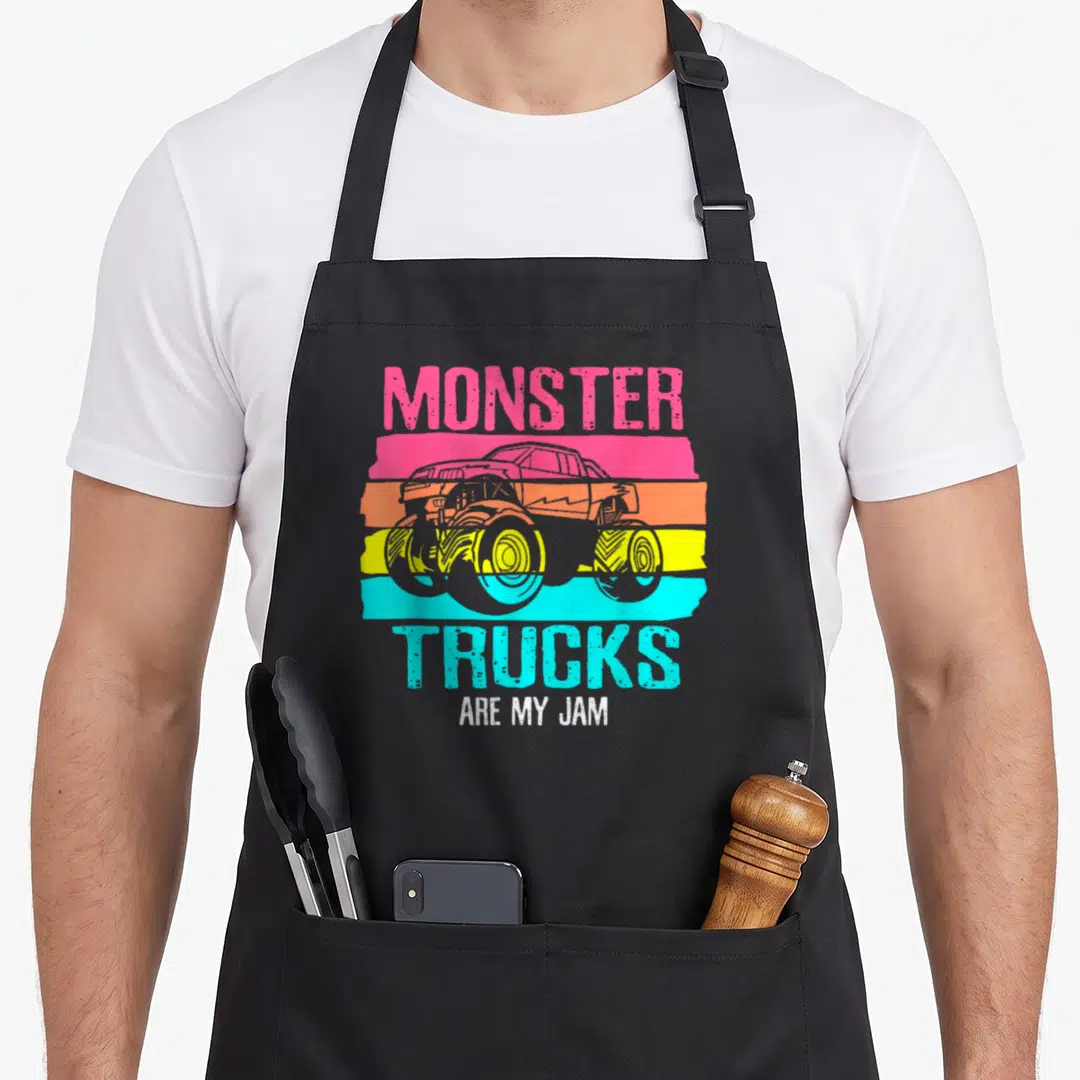 Jam engine truck apron - Retro monster truck on pink orange yellow and cyan stripes with text MONSTER TRUCKS ARE MY JAM — shown on a black cotton canvas apron against a white background