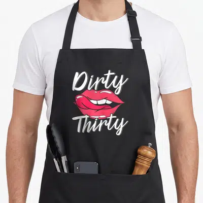 Bday funny dirty aprons - White Dirty Thirty text framing a pair of open pink lips with a tongue sticking out — shown on a black cotton canvas apron against a white background