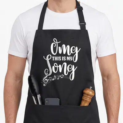 Omg camp songs apron - White stacked Omg THIS IS MY Song text with treble clef and musical notes at bottom left — shown on a black cotton canvas apron against a white background