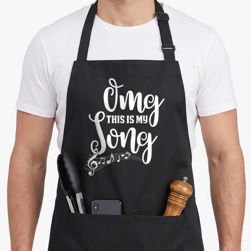Omg camp songs apron - White stacked Omg THIS IS MY Song text with treble clef and musical notes at bottom left — shown on a black cotton canvas apron against a white background