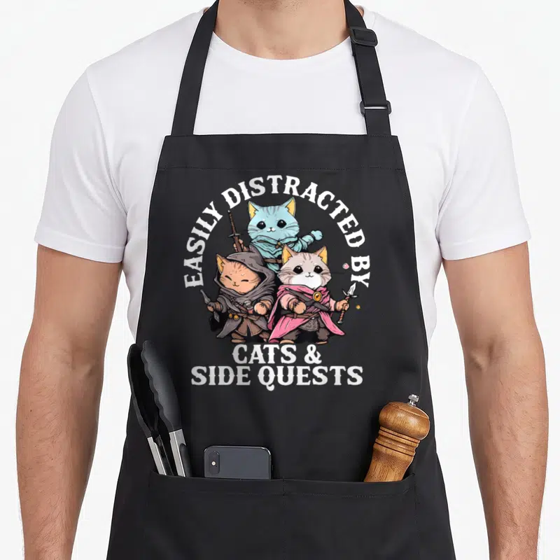 Quest cat dad apron - Three fantasy cats with swords and robes under text easily distracted by cats and side quests — shown on a black cotton canvas apron against a white background