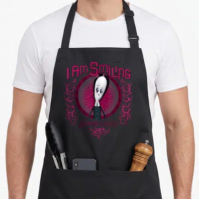Smiling family aprons - Wednesday Addams cartoon portrait with I AM SMILING text and pink swirls — shown on a black cotton canvas apron against a white background
