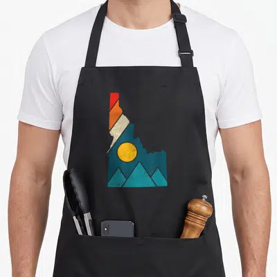 State camping apron - Retro Idaho state silhouette with teal mountains, yellow sun, and red orange cream diagonal — shown on a black cotton canvas apron against a white background