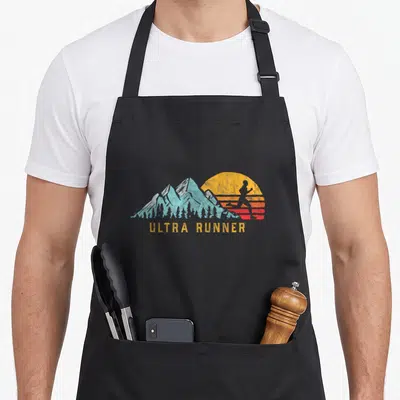 Ultra marathon apron - Retro runner silhouette against sun and mountains with ULTRA RUNNER text in yellow and blue — shown on a black cotton canvas apron against a white background