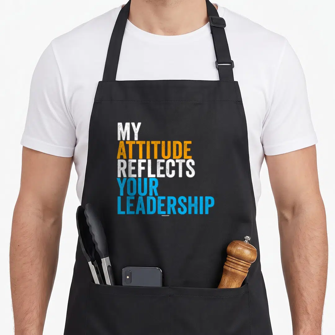 Swagazon attitude aprons - Stacked white orange and blue text reading My Attitude Reflects Your Leadership — shown on a black cotton canvas apron against a white background