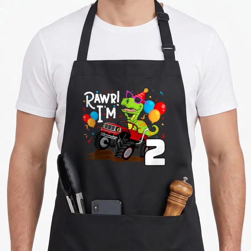 Rex dinosaur truck apron - Green dinosaur in red monster truck with RAWR I'M 2 text and party balloons — shown on a black cotton canvas apron against a white background