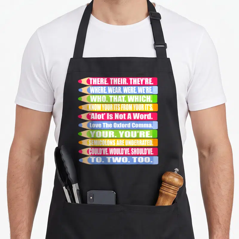 Their spelling of apron - Ten stacked pencils in red blue green yellow white with grammar rules text — shown on a black cotton canvas apron against a white background