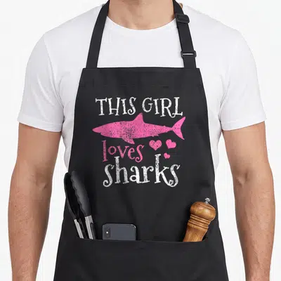 Biologist animal apron - Pink shark silhouette with white text THIS GIRL loves sharks and three hearts — shown on a black cotton canvas apron against a white background