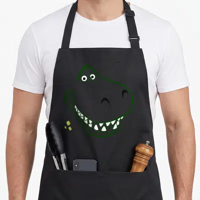 Rex halloween apron - Cartoon dinosaur head with grey skin, green outline, and white teeth on a grey background — shown on a black cotton canvas apron against a white background