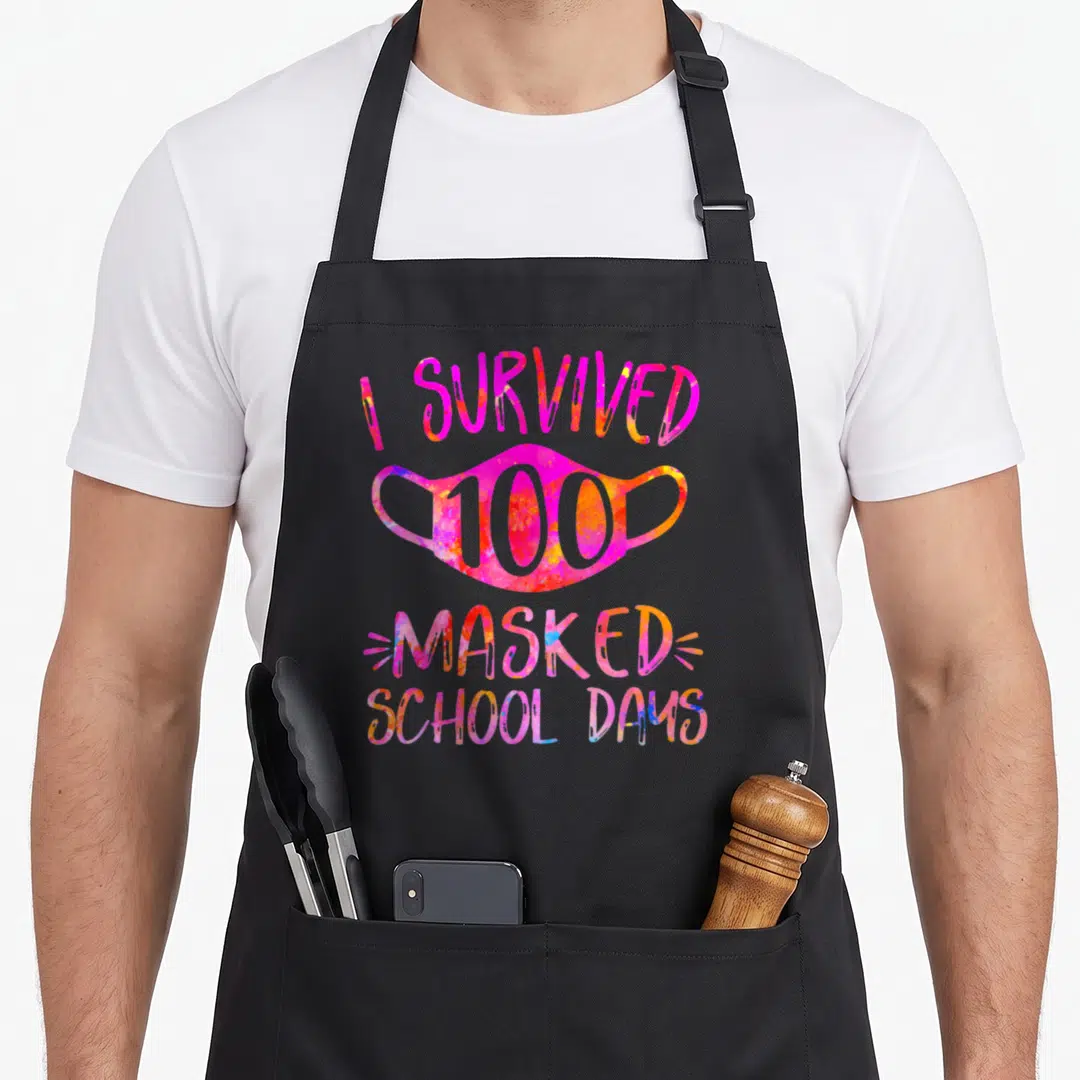 Masked teacher apron - Watercolor face mask with I Survived 100 Masked School Days text in pink and orange — shown on a black cotton canvas apron against a white background