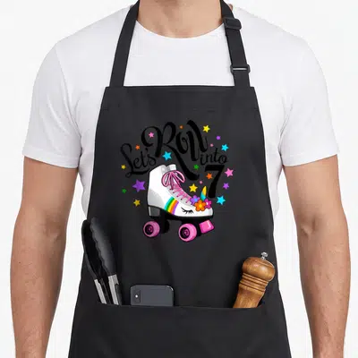 Roll unicorn birthday apron - White roller skate with pink laces, rainbow stripe, unicorn horn, and colorful stars — shown on a black cotton canvas apron against a white background