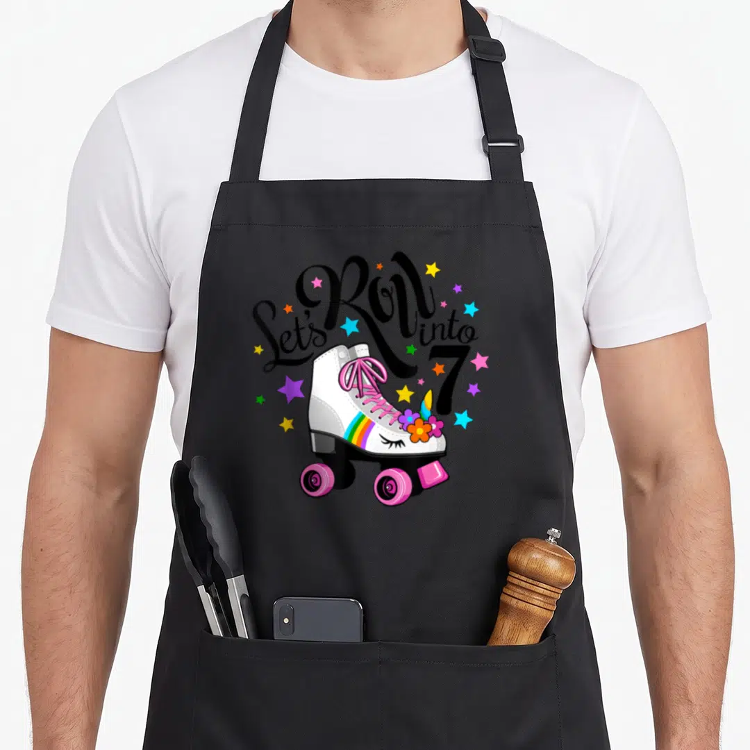 Roll unicorn birthday apron - White roller skate with pink laces, rainbow stripe, unicorn horn, and colorful stars — shown on a black cotton canvas apron against a white background