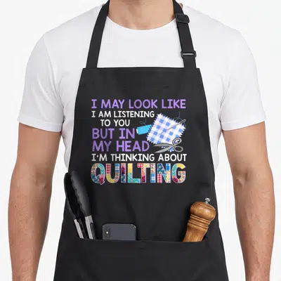 Fabricaholic seamstress apron - Stacked text I MAY LOOK LIKE I AM LISTENING TO YOU BUT IN MY HEAD I'M THINKING ABOUT — shown on a black cotton canvas apron against a white background