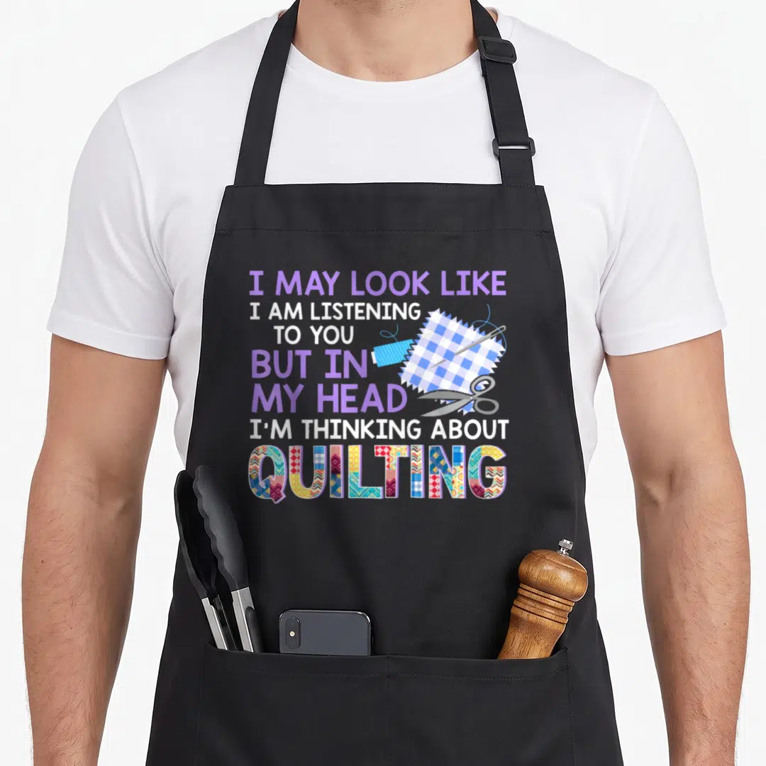 Fabricaholic seamstress apron - Stacked text I MAY LOOK LIKE I AM LISTENING TO YOU BUT IN MY HEAD I'M THINKING ABOUT — shown on a black cotton canvas apron against a white background