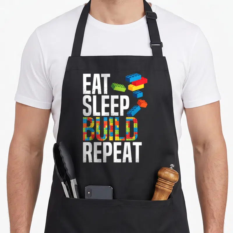 Repeat grill master apron - Stacked text EAT SLEEP BUILD REPEAT with colorful brick patterns filling BUILD and loose — shown on a black cotton canvas apron against a white background