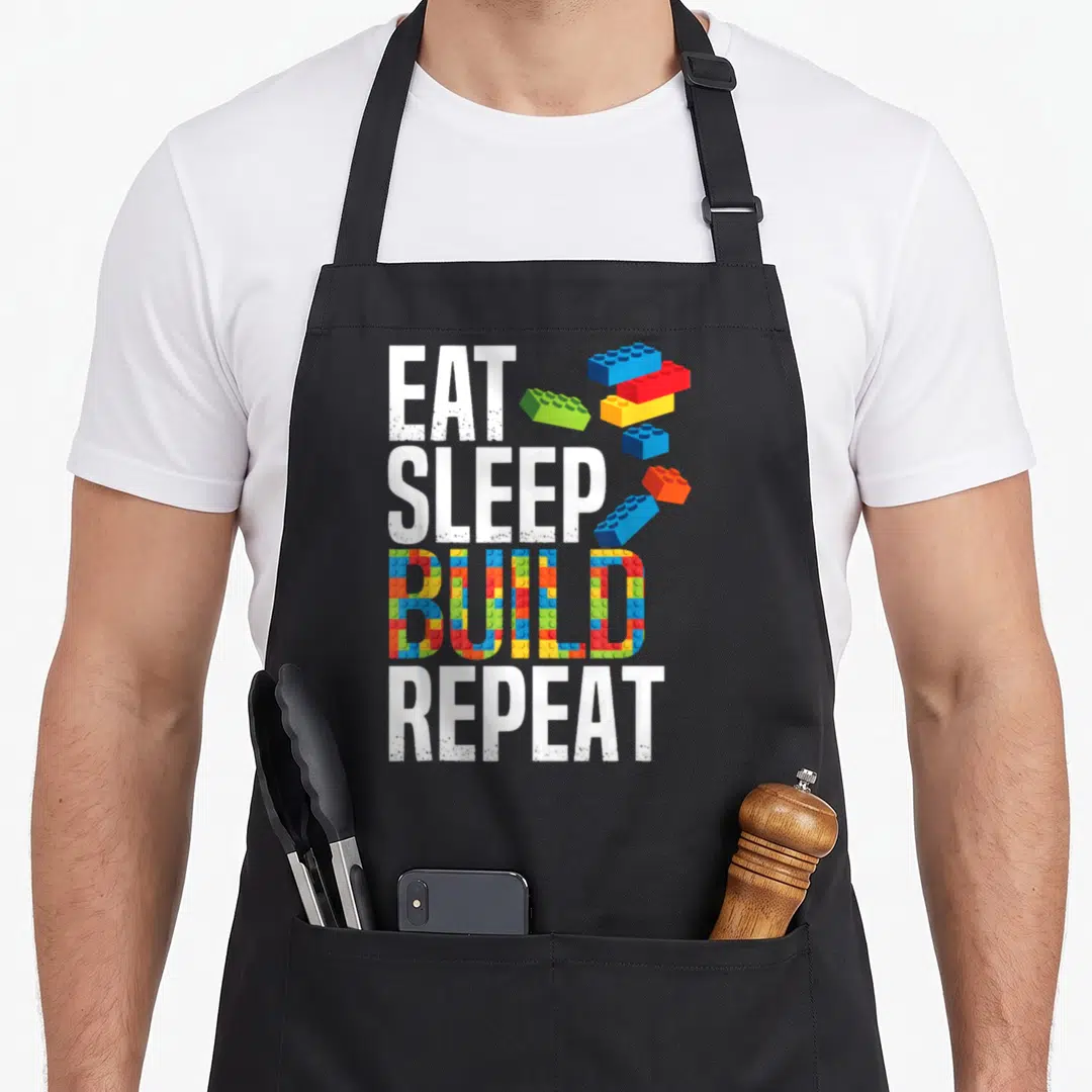 Repeat grill master apron - Stacked text EAT SLEEP BUILD REPEAT with colorful brick patterns filling BUILD and loose — shown on a black cotton canvas apron against a white background