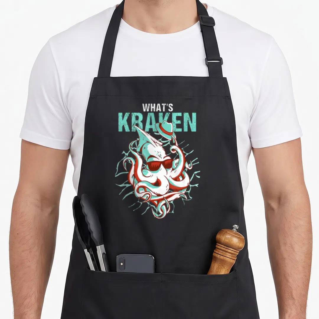 Ocean kraken apron - Cartoon kraken with sunglasses and hat under stacked text reading WHAT'S KRAKEN in teal and white — shown on a black cotton canvas apron against a white background