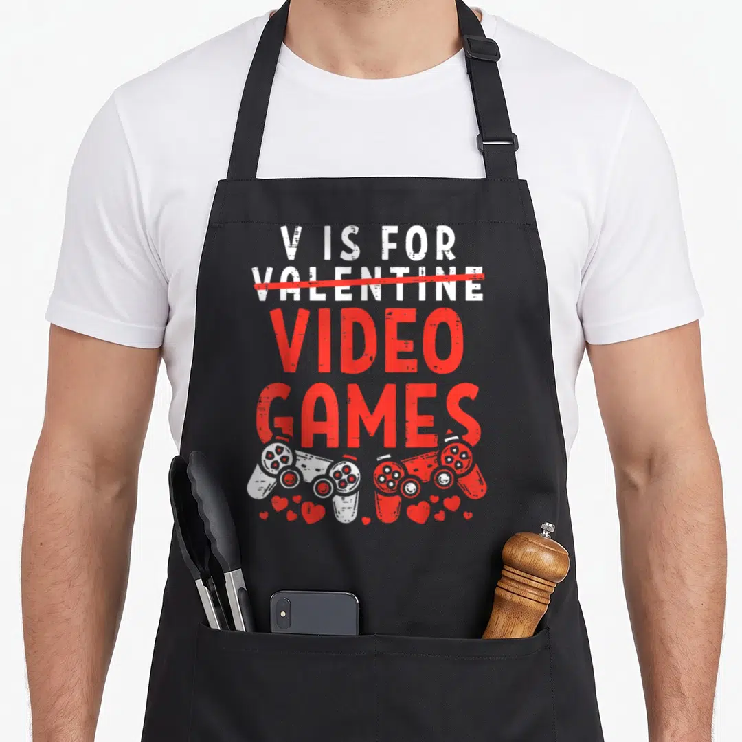 Video valentine apron - V is for Video Games text with red strikethrough and two controllers — shown on a black cotton canvas apron against a white background