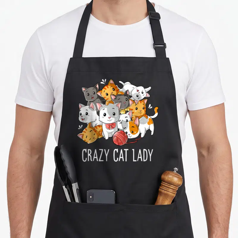 Kitty crazy cat lady apron - Nine cartoon cats in orange white grey and red surrounding a red yarn ball above white text — shown on a black cotton canvas apron against a white background