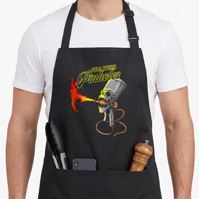 Automotive auto apron - FILL YOUR Pinholes text above skull spray gun breathing fire in grey yellow red orange — shown on a black cotton canvas apron against a white background