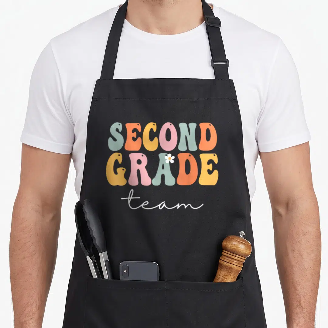 Second love grade apron - Stacked bubbly SECOND GRADE team text in orange yellow pink blue white with small white flower — shown on a black cotton canvas apron against a white background