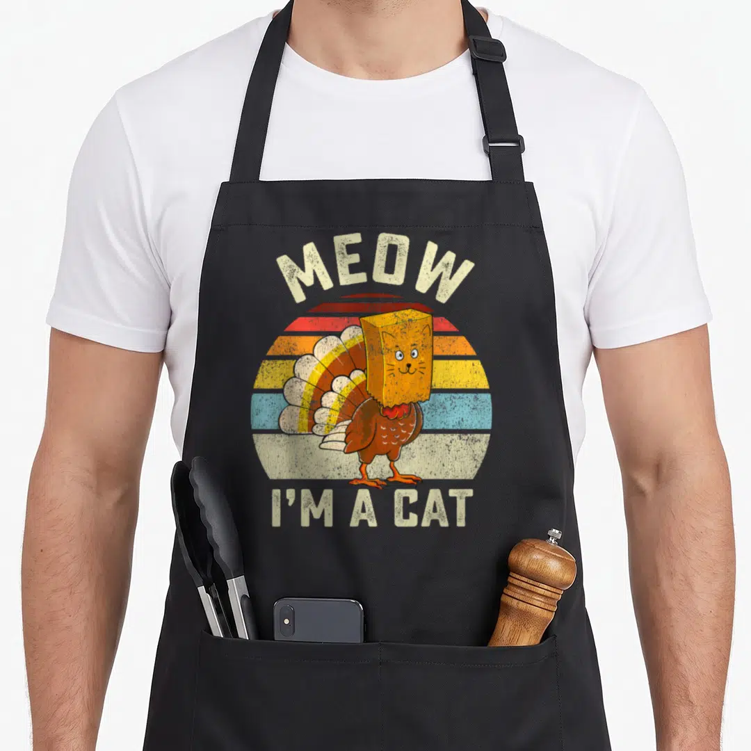 Fake turkey apron - Retro cartoon turkey wearing orange paper bag cat mask in front of striped sun with MEOW I'M A CAT — shown on a black cotton canvas apron against a white background