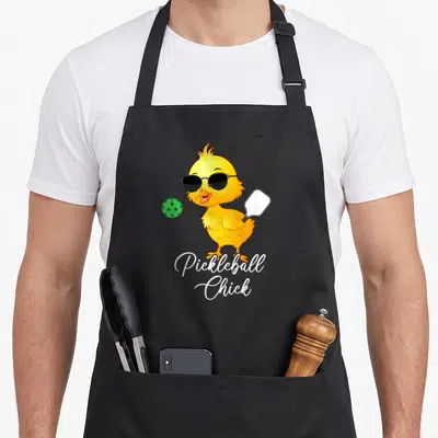 Funny pickleball apron - Cartoon yellow chick with sunglasses holding a paddle next to a green ball above text — shown on a black cotton canvas apron against a white background