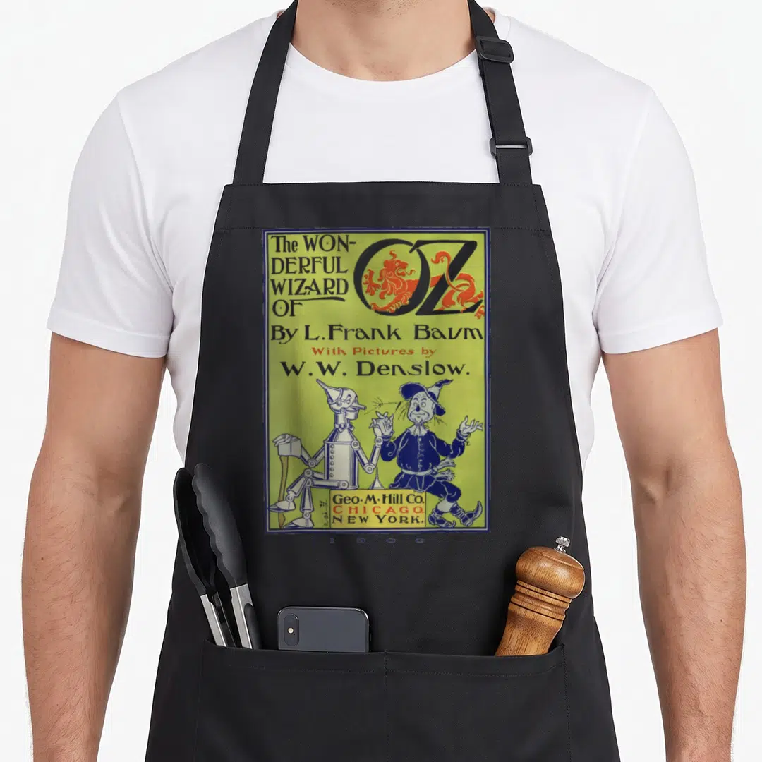 Tin scarecrow apron - 1900 Wizard of Oz book cover print showing Tin Woodman and Scarecrow standing under title text — shown on a black cotton canvas apron against a white background
