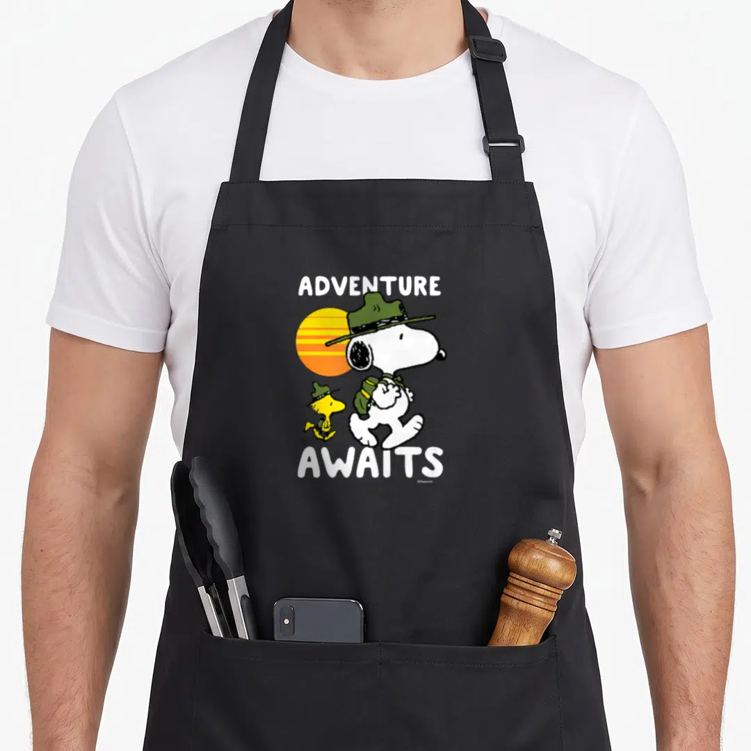 Adventure peanut apron - Snoopy and Woodstock hiking in green hats before a striped orange and yellow sun with white — shown on a black cotton canvas apron against a white background