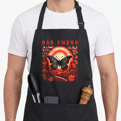 Omen moth apron - BAD OMENS text above black butterfly and red sunburst with floral line art — shown on a black cotton canvas apron against a white background
