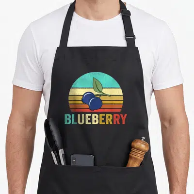 Sunset blueberries apron - Two blueberries and leaf on striped sunset background with BLUEBERRY text — shown on a black cotton canvas apron against a white background