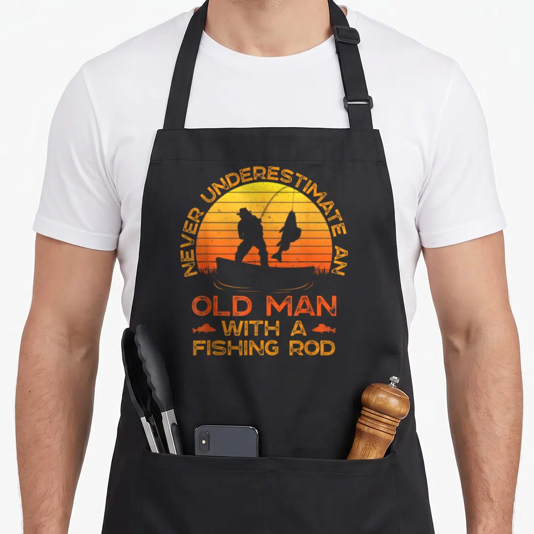 Underestimate fishing aprons - Retro sunset fisherman silhouette with orange text and two small fish icons flanking — shown on a black cotton canvas apron against a white background