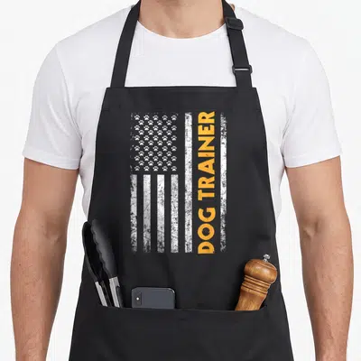 Animal dog trainer apron - Distressed white flag with paw print stars and vertical orange DOG TRAINER text on black — shown on a black cotton canvas apron against a white background