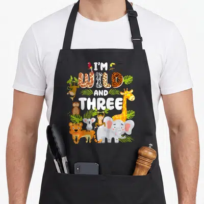Wild three birthday apron - I'M WILD AND THREE text with cartoon tiger, elephant, and birds on black bib — shown on a black cotton canvas apron against a white background