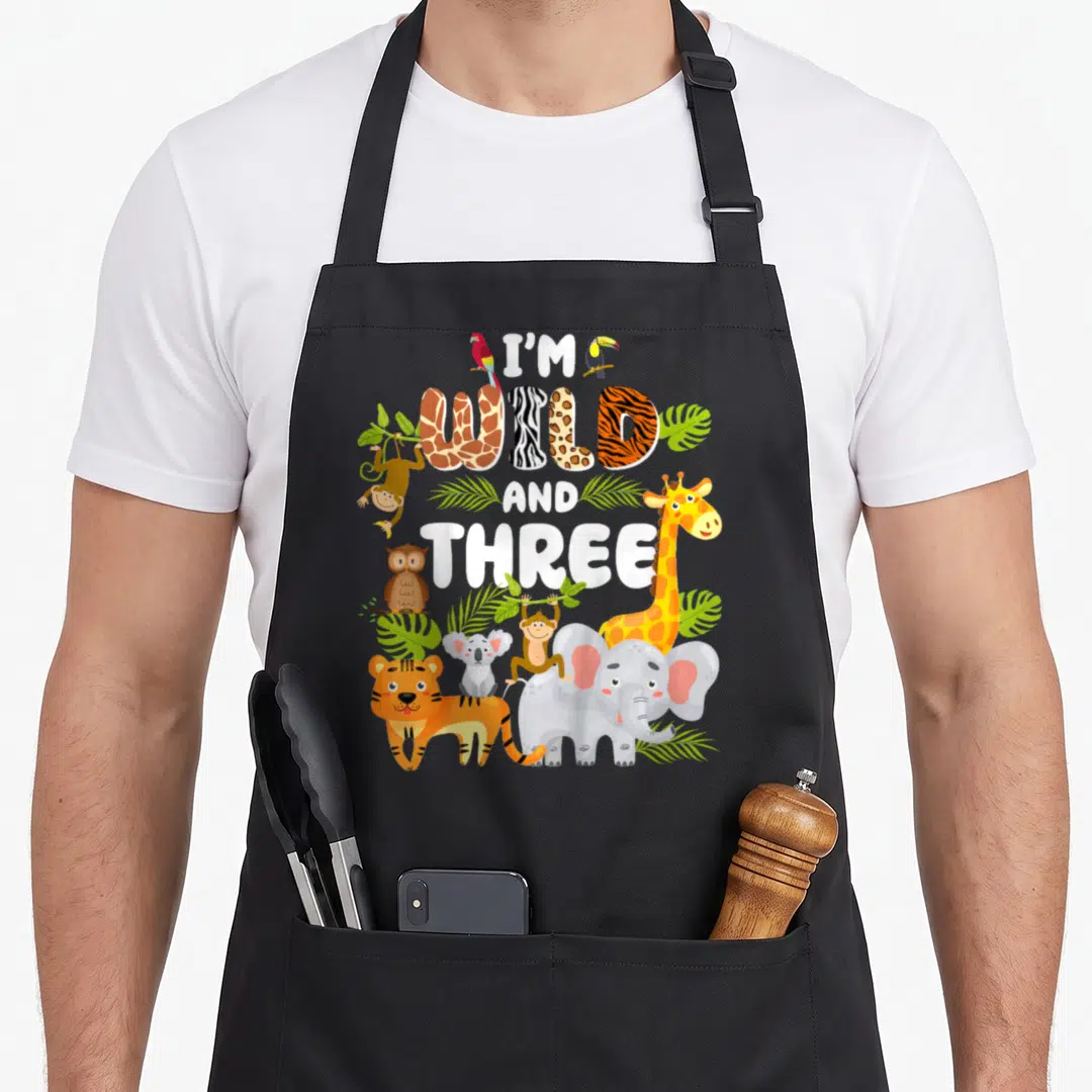 Wild three birthday apron - I'M WILD AND THREE text with cartoon tiger, elephant, and birds on black bib — shown on a black cotton canvas apron against a white background