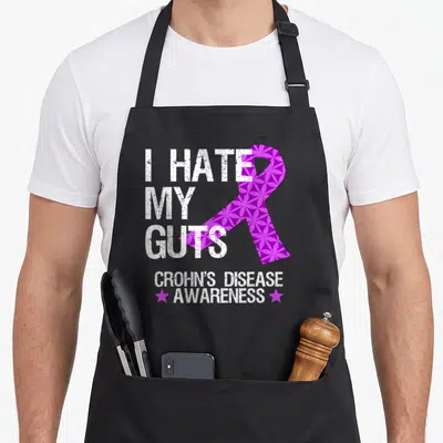 Hate crohns awareness apron - Distressed white text I HATE MY GUTS beside patterned purple ribbon and CROHN'S DISEASE — shown on a black cotton canvas apron against a white background