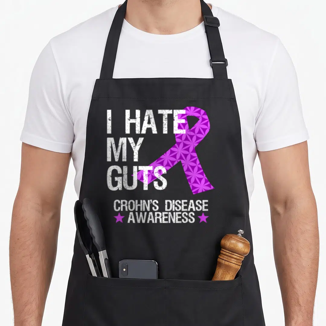 Hate crohns awareness apron - Distressed white text I HATE MY GUTS beside patterned purple ribbon and CROHN'S DISEASE — shown on a black cotton canvas apron against a white background