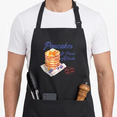 Panic pancake apron - Stack of pancakes with butter, syrup, blueberries, and text Pancakes & Panic Attacks in blue — shown on a black cotton canvas apron against a white background