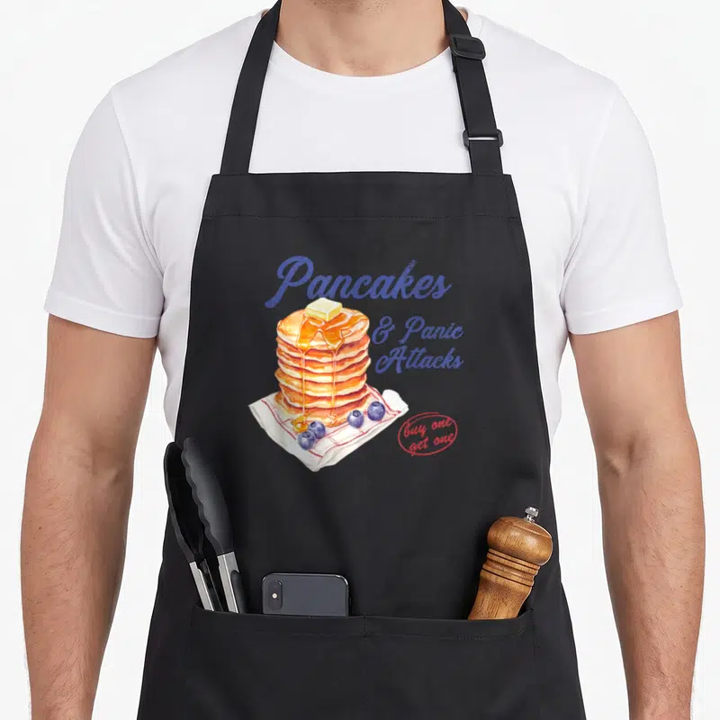 Panic pancake apron - Stack of pancakes with butter, syrup, blueberries, and text Pancakes & Panic Attacks in blue — shown on a black cotton canvas apron against a white background