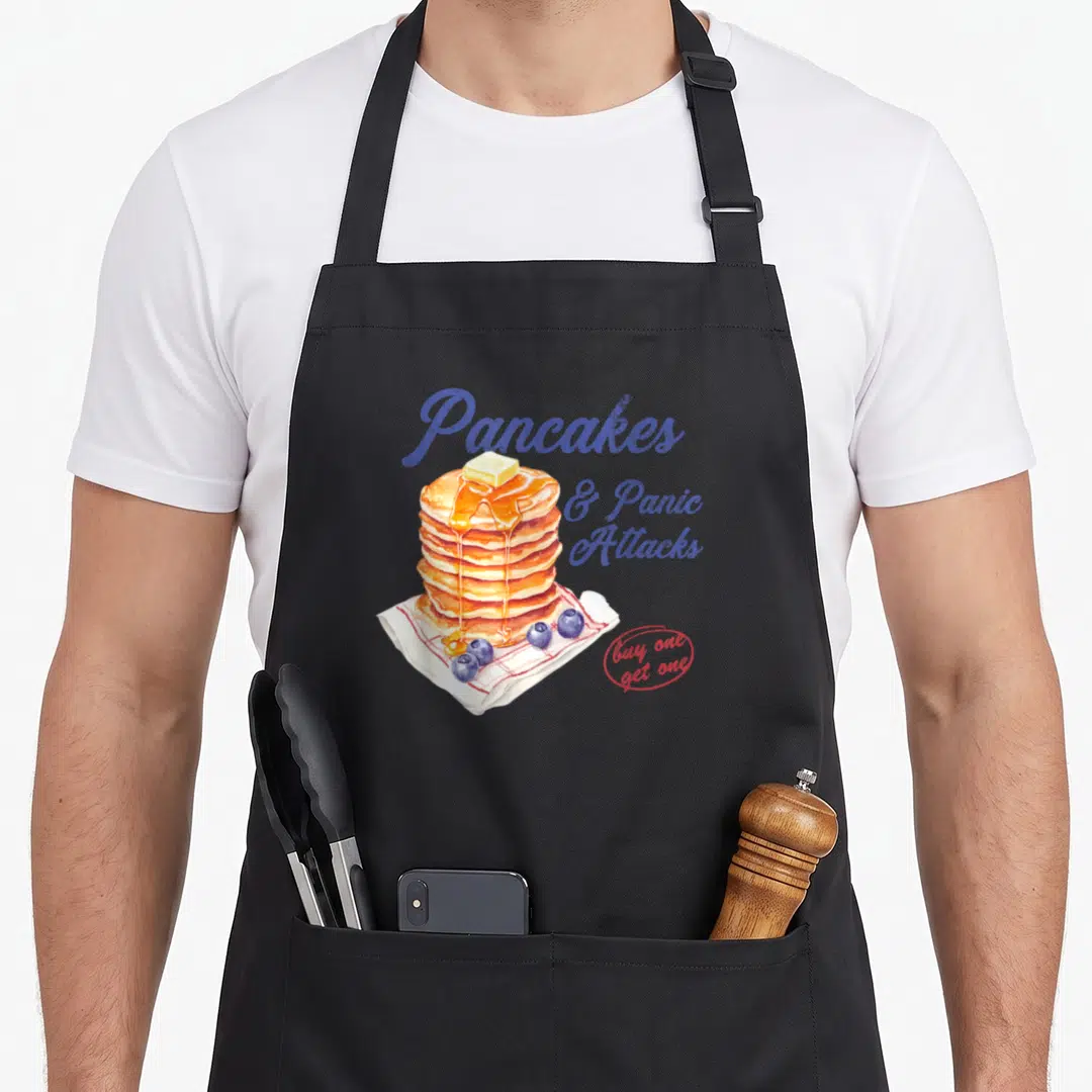 Panic pancake apron - Stack of pancakes with butter, syrup, blueberries, and text Pancakes & Panic Attacks in blue — shown on a black cotton canvas apron against a white background