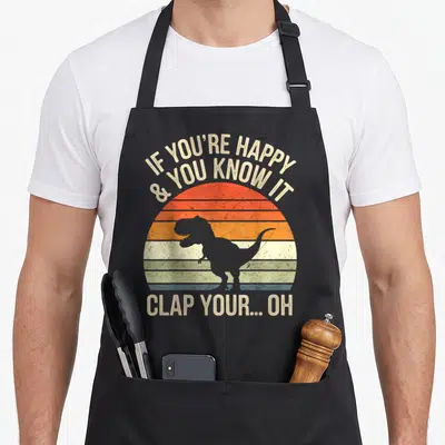 Meme t rex apron - Grey T-Rex silhouette on striped retro sun with clapping nursery rhyme text — shown on a black cotton canvas apron against a white background