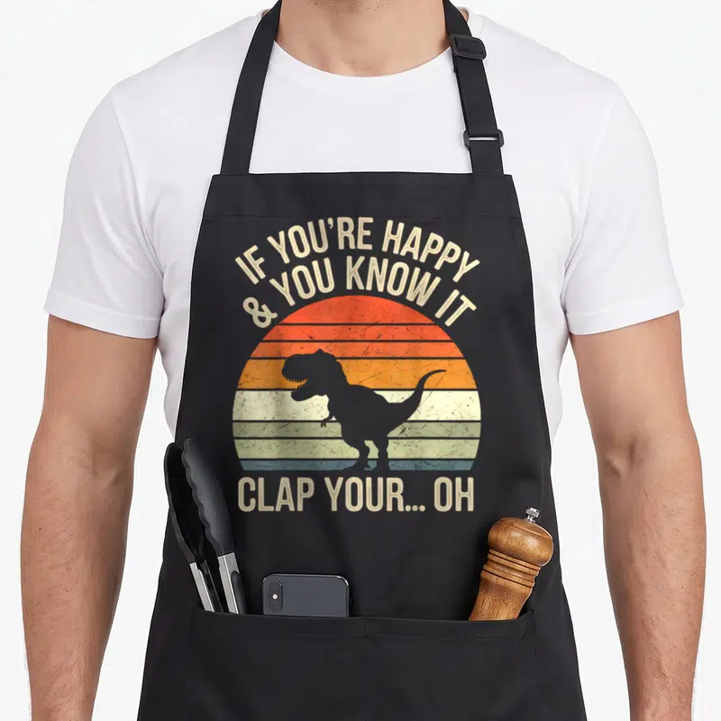 Meme t rex apron - Grey T-Rex silhouette on striped retro sun with clapping nursery rhyme text — shown on a black cotton canvas apron against a white background