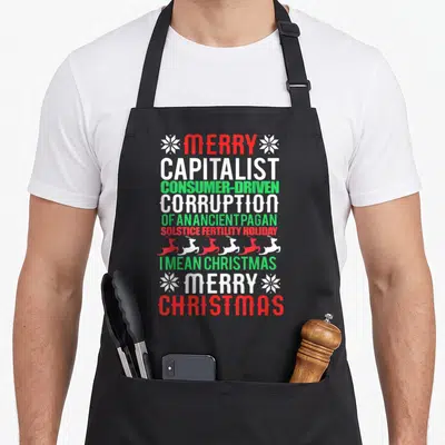 Merry retro christmas aprons - Stacked red white and green text reading Merry Capitalist Consumer Driven Corruption — shown on a black cotton canvas apron against a white background